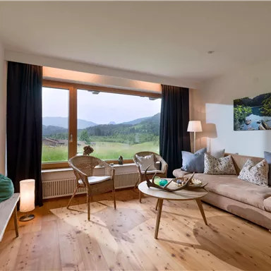 Ein gemütliches Wohnzimmer mit großen Fenstern und Blick auf eine grüne Landschaft. Die Einrichtung ist modern und einladend mit bequemen Möbeln und warmen Holzböden.