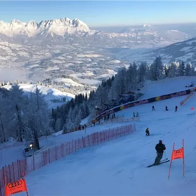 Eine verschneite Skistation mit Pisten und Skifahrern. Im Hintergrund sind beeindruckende Berge und ein klarer Himmel zu sehen.
