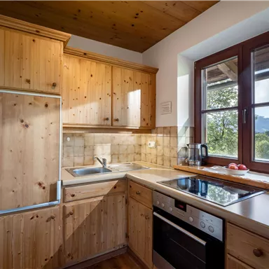 A modern kitchen with wooden cabinets and a bright color scheme. It is equipped with a stove, a sink, and a refrigerator that let light in through the windows.
