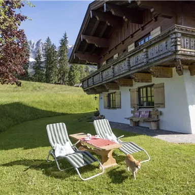 A cozy chalet in a green landscape, surrounded by trees and mountains. In front of the house are two loungers and a table with breakfast.