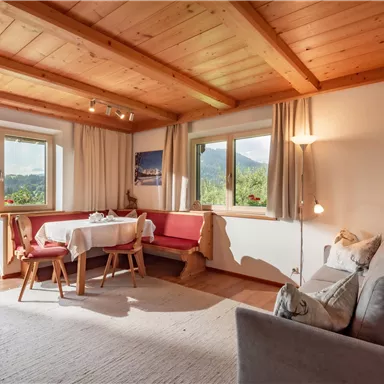 A cozy living room with a wooden beam ceiling and large windows. There is a sofa, a table, and a beautiful view of the surroundings.