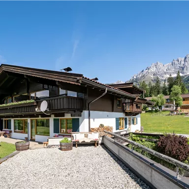 Ein traditionelles Chalet in einer malerischen Landschaft mit Bergen im Hintergrund. Der Garten ist gepflegt und die Sonne scheint klar am blauen Himmel.