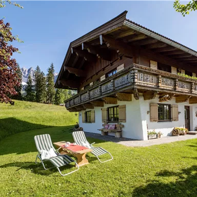 A cozy alpine house with a balcony and a well-kept garden. In the foreground, there are loungers on the green meadow.