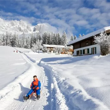 Ein Junge rodelt auf einem schneebedeckten Weg. Im Hintergrund sind verschneite Bäume und ein gemütliches Haus zu sehen.