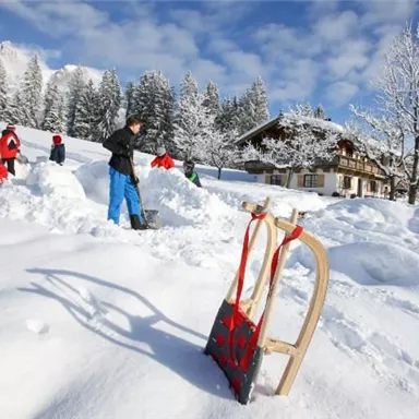 A snowy landscape with several people shoveling snow. In the foreground, there is a sled and in the background, you can see a wooden house and snow-covered trees.