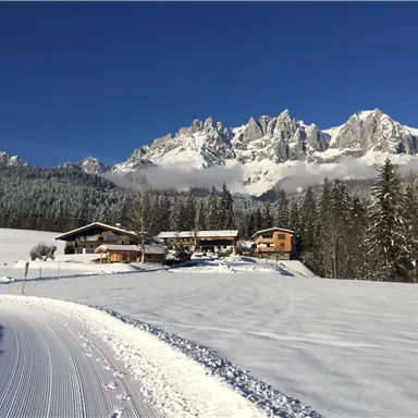 Eine malerische Winterlandschaft mit schneebedeckten Hügeln und majestätischen Bergen im Hintergrund. Hütten stehen in einer ruhigen Umgebung umgeben von Bäumen.