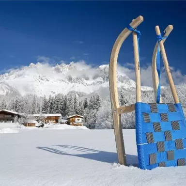 Ein rodeliger Schlitten steht im Schnee vor einem malerischen Bergpanorama. Im Hintergrund sind verschneite Bäume und traditionelle Hütten sichtbar.