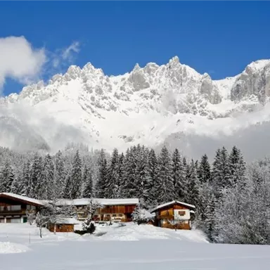 Eine malerische Winterlandschaft mit schneebedeckten Bergen im Hintergrund. Vorne sind gemütliche Holzhäuser umgeben von verschneiten Bäumen.