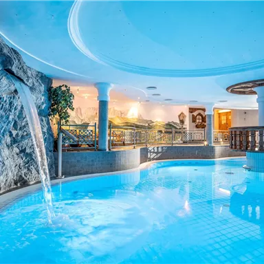 A modern swimming pool with clear water and a waterfall. The surroundings are elegantly designed, with plants and tasteful lighting.