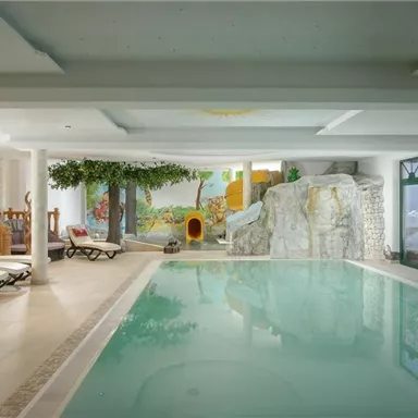 Ein schöner Innenpool mit entspannenden Liegen und einer Dekoration aus Felsen und Pflanzen. Helle Fenster lassen viel Tageslicht herein.