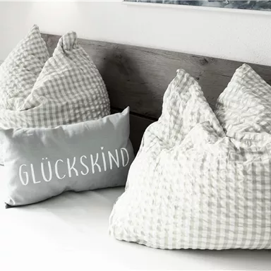Ein gemütliches Bett mit Kissen in grün-weißem Karomuster. Ein Kissen mit der Aufschrift „GLÜCKSKIND“ ergänzt die dekorative Atmosphäre.