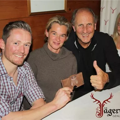 Eine Gruppe von vier Personen sitzt an einem Tisch in einem gemütlichen Raum. Alle lächeln und zeigen Freude, während einer von ihnen ein kleines Geschenk präsentiert.