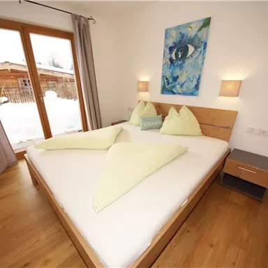 Ein modernes Schlafzimmer mit einem großen Bett und holzfarbenem Boden. Die Wände sind hell, und es gibt ein Kunstwerk sowie Fenster mit Blick auf eine Außenansicht.