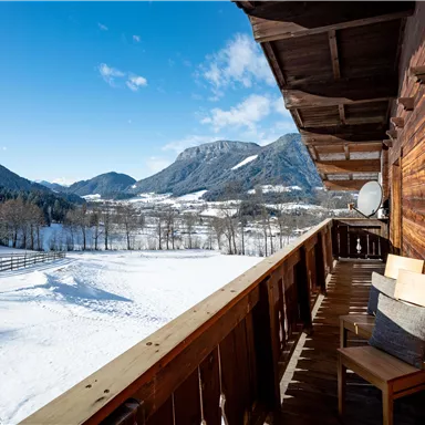 A balcony with wooden cladding and two chairs overlooks a snow-covered landscape. In the background, majestic mountains rise under a clear blue sky.