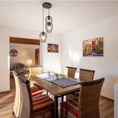 A cozy dining area with a wooden table and rattan-like chairs. Some pictures hang on the walls, and a plant sits on the table.