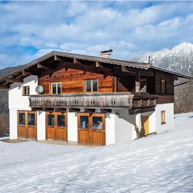 A traditional wooden house in the Alps stands against a backdrop of snow-covered mountains. The sky is partially cloudy, creating a calm winter atmosphere.
