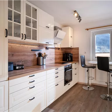 A modern kitchen with white cabinets and wood surfaces. There is a small dining area with high chairs and plenty of natural light.
