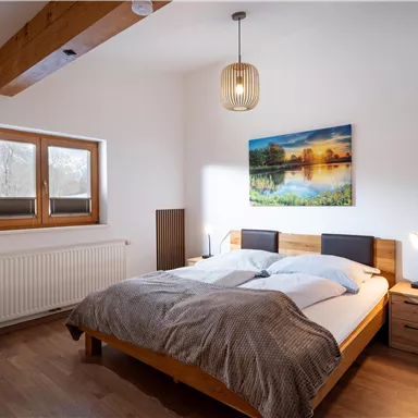 A cozy bedroom with a large bed and a wooden stand. A picture of a landscape hangs on the wall, and the light falls through the windows.