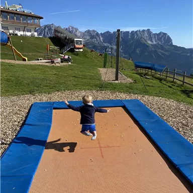 Ein kleines Kind springt fröhlich auf einem Trampolin in den Bergen. Im Hintergrund sind die majestätischen Berge und ein klarer blauer Himmel zu sehen.