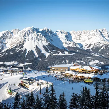 Eine schöne Winterlandschaft mit schneebedeckten Bergen. Im Vordergrund sieht man ein Skigebiet mit mehreren Skiliften und einer Hütte.