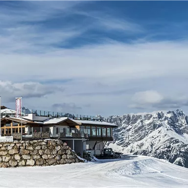 Eine Berghütte in einer verschneiten Landschaft mit beeindruckenden Bergen im Hintergrund. Der Himmel ist klar und strahlend blau.