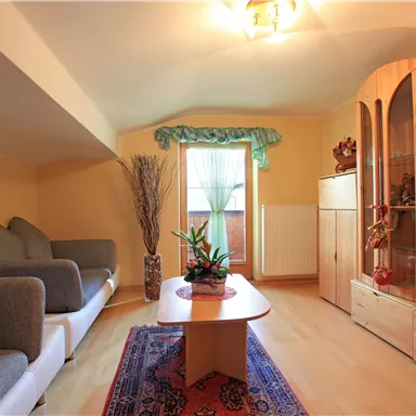 A cozy living room with two gray sofas and a wooden table. A plant stands by a window, and a television is placed in the room.