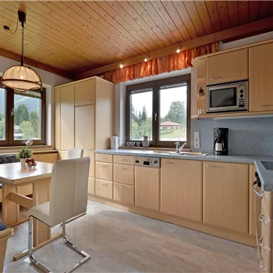 A modern kitchen with a wooden ceiling and large windows. The dining area has a table and chairs, as well as a kitchenette with various appliances.