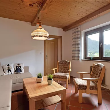 A cozy living room with a light sofa and two wooden chairs. Large windows provide a view of the landscape.