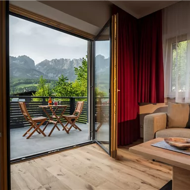 A cozy room with a view of the mountains. Outside is a balcony with wooden chairs and a table.