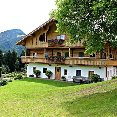 A traditional alpine house with wooden cladding and a balcony. Surrounded by a green garden and mountains in the background.