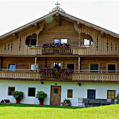 A traditional alpine wooden house with a balcony and colorful flowers. It stands in a green setting, surrounded by trees.