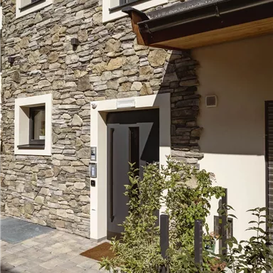 Ein modernes Haus mit einer Steinfassade und großen Fenstern. Der Eingangsbereich ist mit Pflanzen geschmückt.