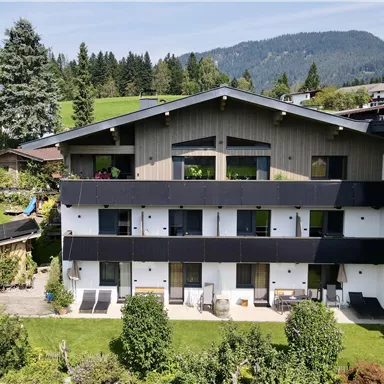 A modern building surrounded by green meadows and trees. In the background, mountains and a blue sky can be seen.