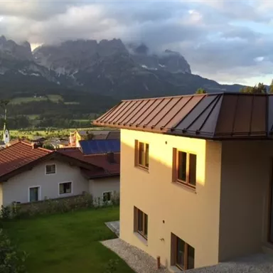 Ein modernes Haus mit einem schönen Garten, umgeben von Bergen. Im Hintergrund sind weitere Häuser und eine malerische Landschaft zu sehen.