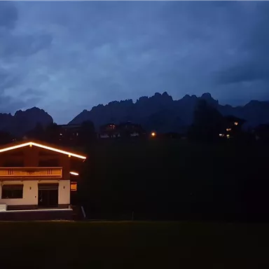 Ein beleuchtetes Chalet in den Bergen bei Dämmerung. Im Hintergrund erheben sich die schroffen Gipfel.