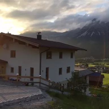 Ein modernes Haus in einer malerischen Berglandschaft. Die Sonne scheint hinter den Bergen und sorgt für eine gemütliche Atmosphäre.