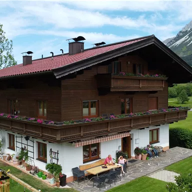 Ein traditionelles bayerisches Haus mit Holzverkleidung und einem roten Dach. Im Hintergrund sind grüne Wiesen und hohe Berge zu sehen.