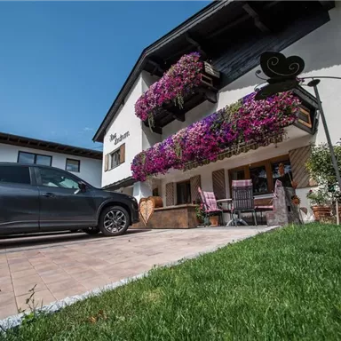 Ein gemütliches Haus mit bunten Blumenampeln und einer schönen Terrasse. Vor dem Haus steht ein graues Auto auf einem Pflasterweg.