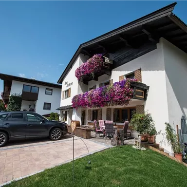 Ein schönes Haus mit lila Blumenbalkonen und einer gepflegten Außenanlage. Vor dem Gebäude stehen ein Auto und Gartenmöbel.