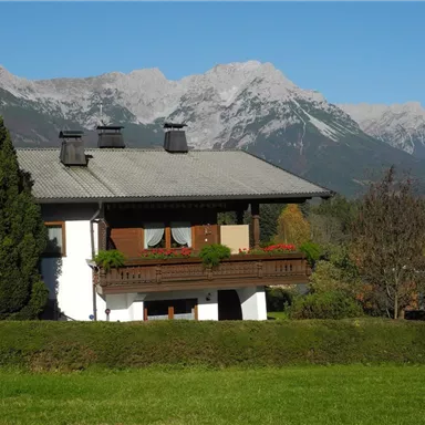 Ein schönes Haus mit Balkon in einer ländlichen Umgebung. Im Hintergrund sind beeindruckende Berge und ein klarer blauer Himmel zu sehen.