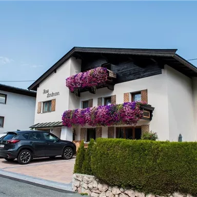 Ein modernes Haus mit bunten Blumenbalkonen. Vor dem Haus stehen zwei Autos auf einem gepflasterten Parkplatz.