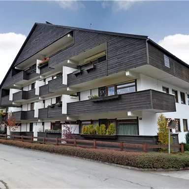 A modern residential building with dark wood design and white walls. The surroundings are green with shrubs and trees.
