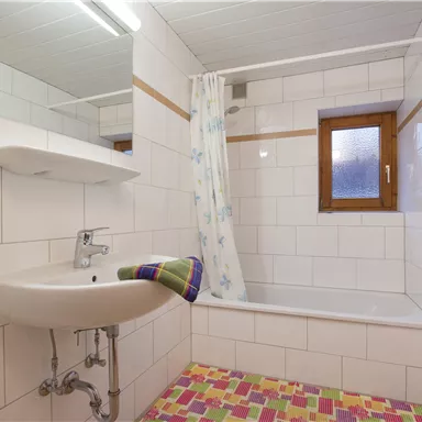 A simple bathroom with a tub, sink, and a window. The walls are tiled and the floor has a colorful pattern.
