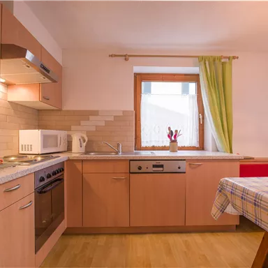 A modern kitchen with wooden cabinets and a large window. The room is bright and friendly, with a table and chairs.