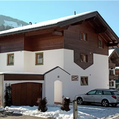 A modern house with wooden cladding, surrounded by snow. In the foreground is a parking lot with a car.