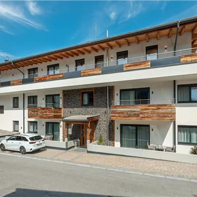 A modern building with multiple floors and wooden cladding. In front, there is a bright car and the surroundings are peaceful with a view of the mountains.