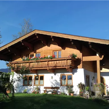Ein traditionelles Alpenhaus mit Holzverkleidung und einem großen Balkon. Umgeben von Grünflächen und einem klaren blauen Himmel.