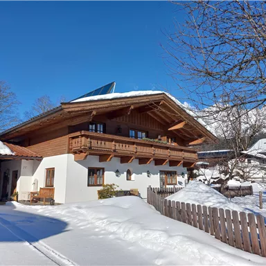 Ein traditionelles Chalet im Winter mit schneebedecktem Grundstück. Der blaue Himmel und die umliegenden Bäume verleihen der Szenerie eine ruhige Atmosphäre.