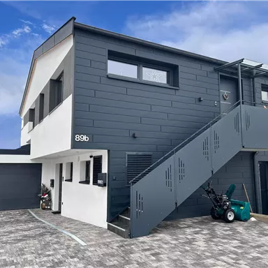 Ein modernes, zweigeschossiges Gebäude mit einer grauen Fassade und einer breiten Außentreppe. Vor dem Gebäude befindet sich ein gepflasterter Parkplatz mit Geräten zur Gartenpflege.