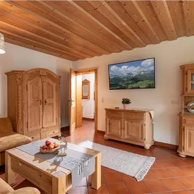 A cozy living space with wood paneling and a bright decor. A couch, a table with fruit, and a TV screen in the background are visible.